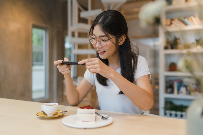 Seorang wanita sedang memfoto dessert kue stroberi dan kopi yang disajikan di depannya.