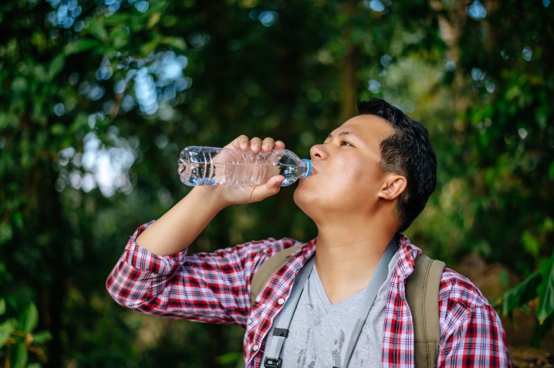 Seorang pemuda asia sedang meminum air mineral dari botol di tengah taman