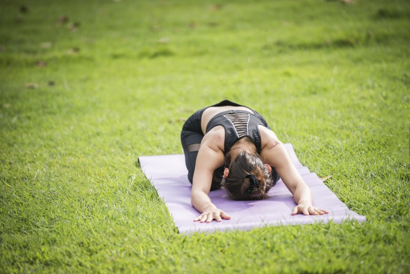 Seorang wanita sedang melakukan yoga dengan pose child  di atas mat berwarna ungu di luar ruangan.